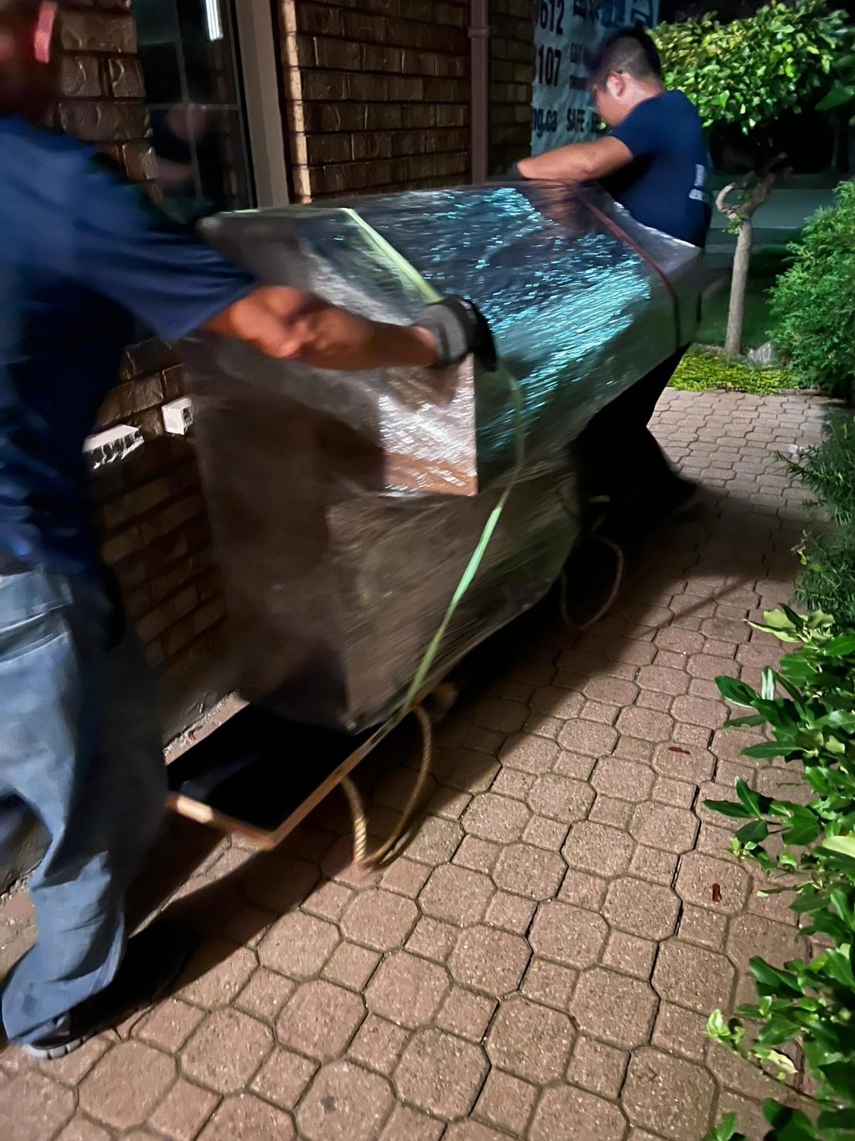 Movers wheeling a wrapped upright piano on a skid board along a walkway to the truck
